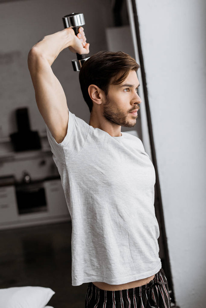 handsome young man in pajamas exercising with dumbbell and looking away in the morning - Foto, Bild