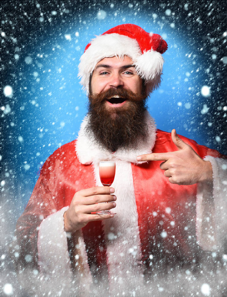 handsome bearded santa claus man with long beard on smiling face holding glass of alcoholic shot in red christmas or xmas sweater and new year hat on blue studio background - Фото, изображение