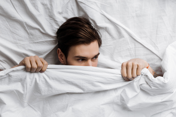 top view of handsome young man hiding under blanket in bed at home and looking away - Фото, изображение
