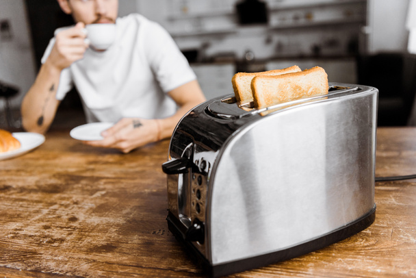Genç adam ön plan üzerinde ekmek kızartma makinesi ile evde kahve içmek kadeh kırpılmış - Fotoğraf, Görsel