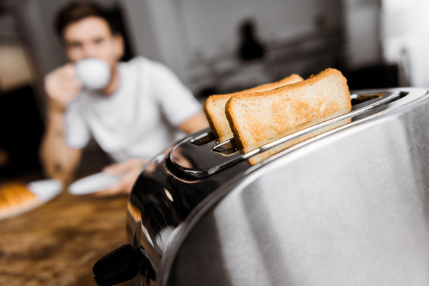 young man drinking coffee at home with toaster on foreground - Foto, afbeelding