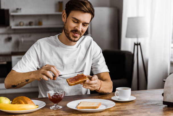 happy young man applying jam onto toast at home - Fotó, kép