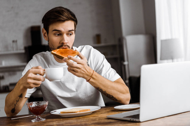 συναισθηματική νέος ελεύθερος επαγγελματίας τρώει τοστ με μαρμελάδα και καφέ και κοιτάζοντας οθόνη φορητού υπολογιστή στο σπίτι - Φωτογραφία, εικόνα