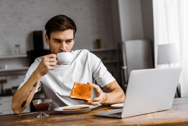ハンサムな若いフリーランス ジャムとコーヒーとトーストを食べて、家でノート パソコンの画面を見て - 写真・画像