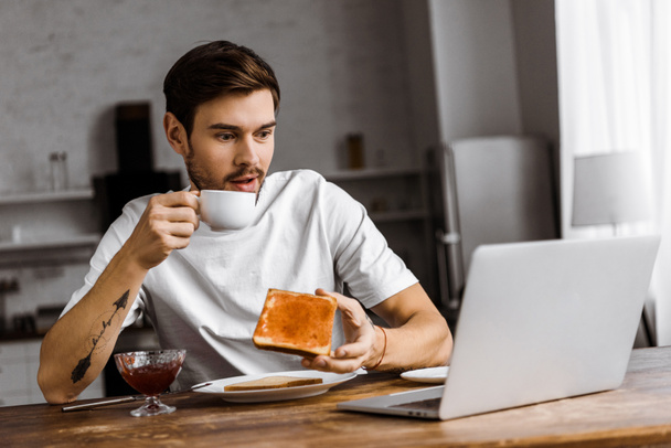 chocado jovem freelancer comer torrada com geleia e café e olhando para tela do laptop em casa
 - Foto, Imagem