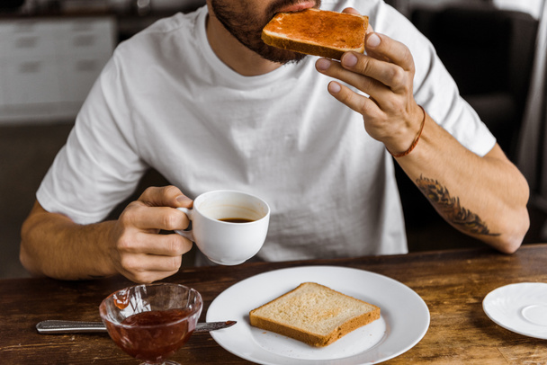 oříznutý snímek mladého muže jíst toasty s džemem a pití kávy doma - Fotografie, Obrázek