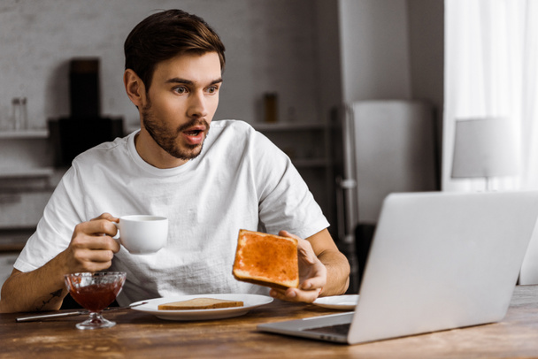 шокований молодий фрілансер їсть тости з джемом і дивиться на екран ноутбука вдома
 - Фото, зображення