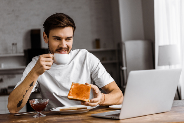 reçelli ekmek yeme ve evde dizüstü bilgisayar ekranına bakarak Genç freelancer yapması - Fotoğraf, Görsel