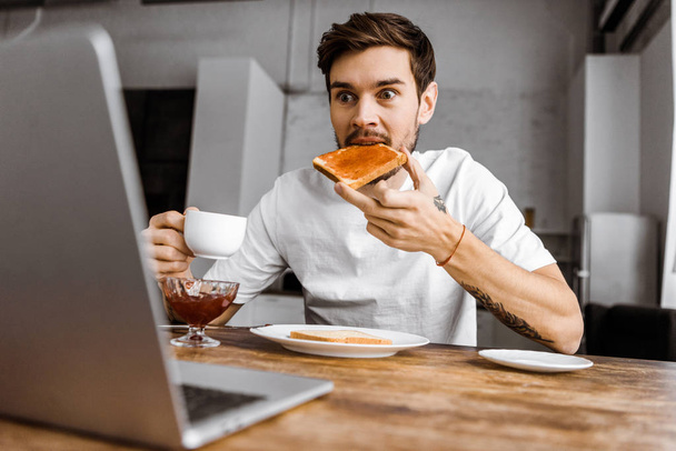 έκπληκτος νέος ελεύθερος επαγγελματίας τρώει τοστ με μαρμελάδα και κοιτάζοντας οθόνη φορητού υπολογιστή στο σπίτι - Φωτογραφία, εικόνα