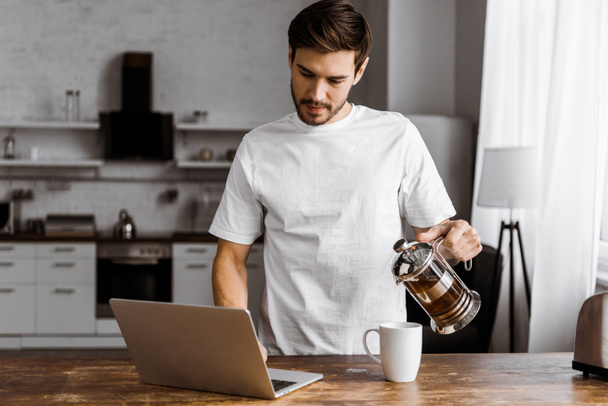 attractive young freelancer with cup of tea, croissant and laptop working on kitchen at home - 写真・画像