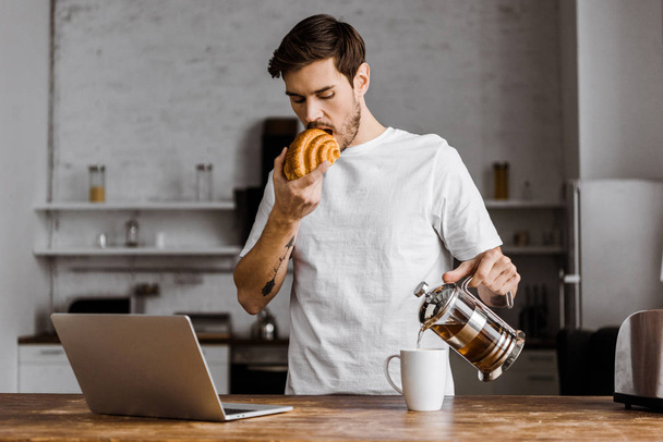 atractivo joven freelancer con taza de té, croissant y portátil trabajando en la cocina en casa
 - Foto, Imagen