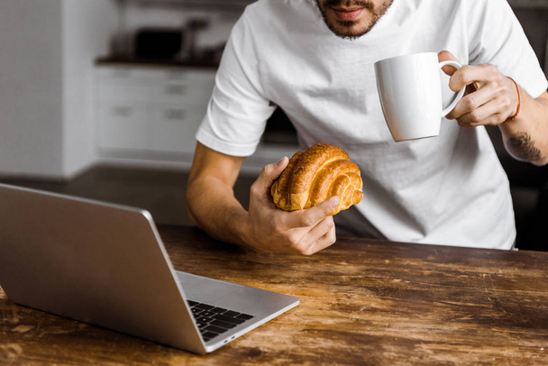 rajattu laukaus houkutteleva nuori freelancer kupin teetä, croissant ja kannettava tietokone työskentelee keittiössä kotona
 - Valokuva, kuva