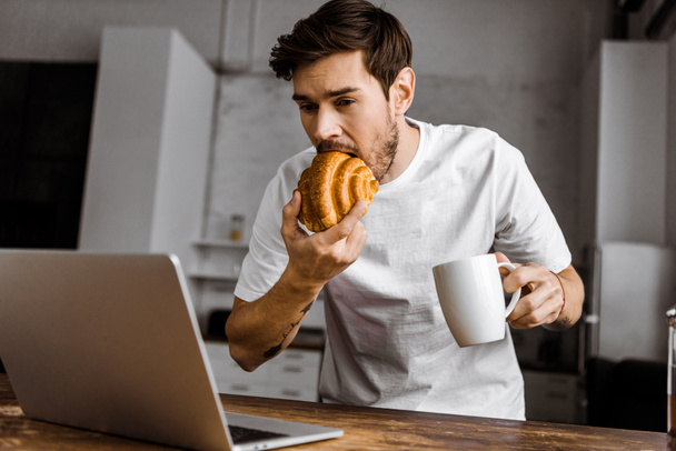 hungrige junge Freiberuflerin mit Tee, Croissant und Laptop in der heimischen Küche - Foto, Bild