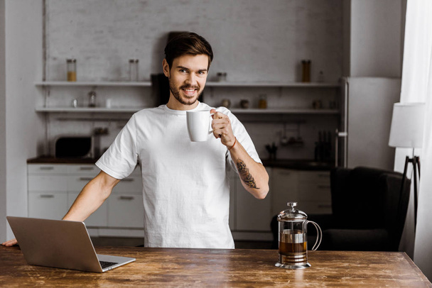 handsome young freelancer with cup of tea and laptop working on kitchen at home - Valokuva, kuva