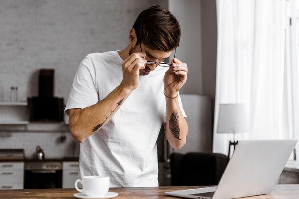 atractivo joven freelancer en gafas con café y portátil en la cocina en casa
 - Foto, Imagen