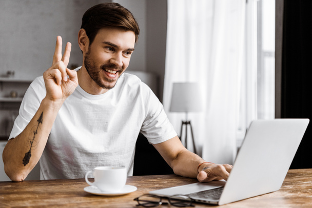 handsome young man with coffee making video call with laptop and making peace gesture at home - Photo, Image