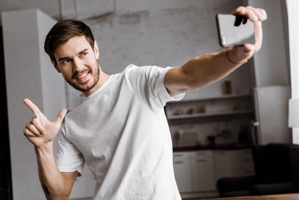 handsome young man taking selfie and making gun gesture at home - Фото, зображення