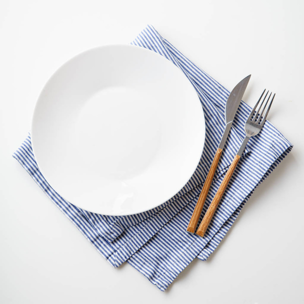 White empty plate with knife and fork on striped blue and white textile napkin isolated on white background, close-up  - Zdjęcie, obraz