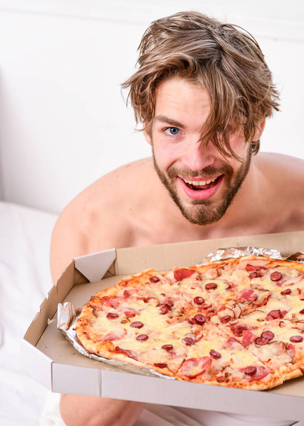 Man likes pizza for breakfast. Sexy man eat pizza lying on bed. Portrait of lazy muscular man eating pizza while laying on a bed at home. Home pizza. - Photo, Image