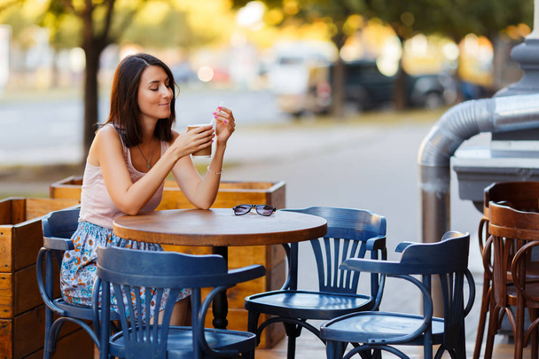 Retrato de una hermosa dama feliz sonriente con un café caliente sentado en una terraza de verano en un café. Una joven está sentada y bebiendo café en la mesa de un restaurante
. - Foto, imagen