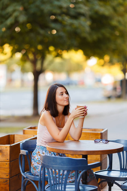 Портрет красивой улыбающейся счастливой леди с горячим кофе, сидящей на летней террасе в кафе. Молодая девушка сидит и пьет кофе за столом ресторана
. - Фото, изображение