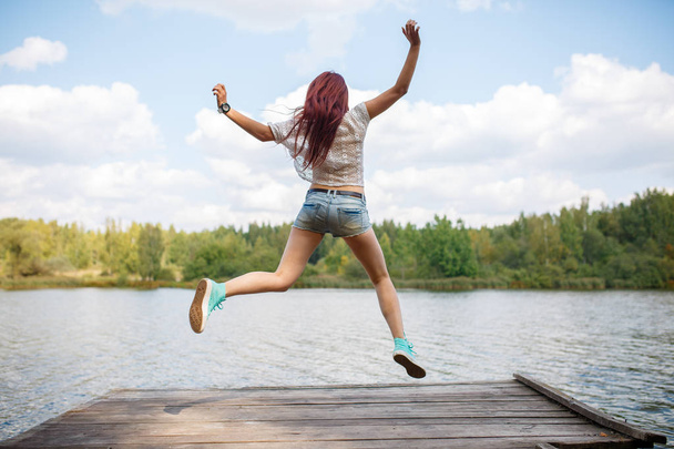 Photo from back of young jumping woman on wooden bridge by river - Photo, image