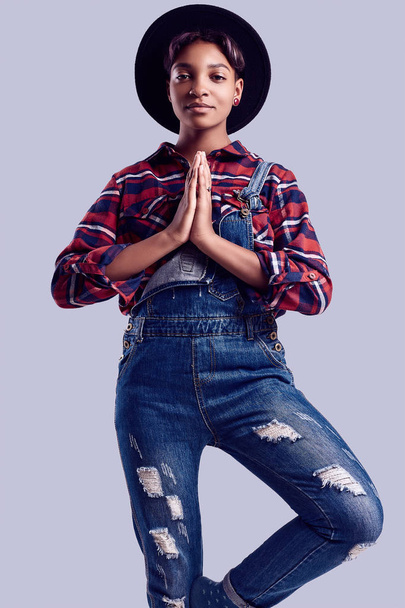 Sinnliches Porträt von Glamour eleganten schwarzen Hipster-Mädchen mit kurzen Haaren in einem karierten Hemd, Jeans-Overalls und Modehut posiert auf buntem Hintergrund im Studio - Foto, Bild