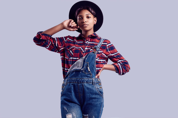 Sensual portrait of glamor elegant black hipster girl with short hair in a plaid shirt, denim overalls and fashion hat posing on colorful background in studio - Photo, Image