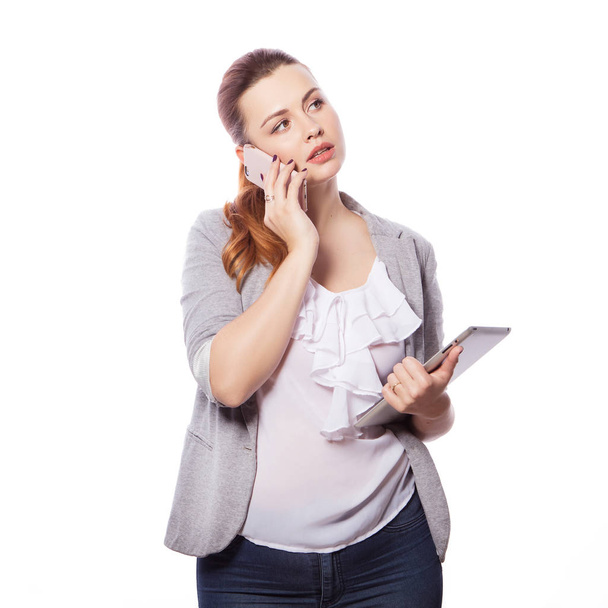 Γυναίκα μελαχρινή συν μέγεθος σε smart casual ντύσιμο (jacker και μπλούζα) σε ένα απομονωμένο άσπρο φόντο. Κατέχει gadgets στα χέρια ger: tablet και τηλέφωνο. Ανεξάρτητος επιχειρηματική ιδέα: εργασία στο διαδίκτυο, Smm - Φωτογραφία, εικόνα