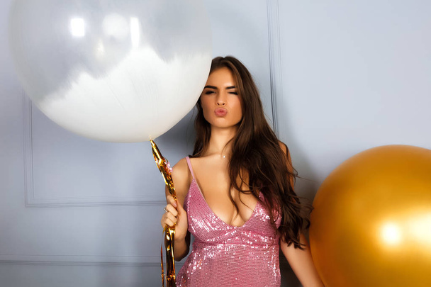 Γοητευτική νεαρή γυναίκα σε φανταχτερό ροζ φόρεμα διασκεδάζοντας με δύο μπαλόνια στο πάρτι. Γιορτάστε μια εκδήλωση. Γίνει φωτεινό μακιγιάζ και χτένισμα. Κρατώντας το μεγάλο λευκό μπαλόνι με χρυσές ρίγες. Φιλιά πρόσωπο - Φωτογραφία, εικόνα