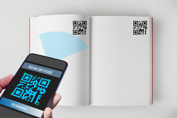 Ruka držící smartphone s skenování Qr kód na obrazovce otevřené knihy na bílém pozadí, inteligentní technologie koncept a Qr kódy idea - Fotografie, Obrázek