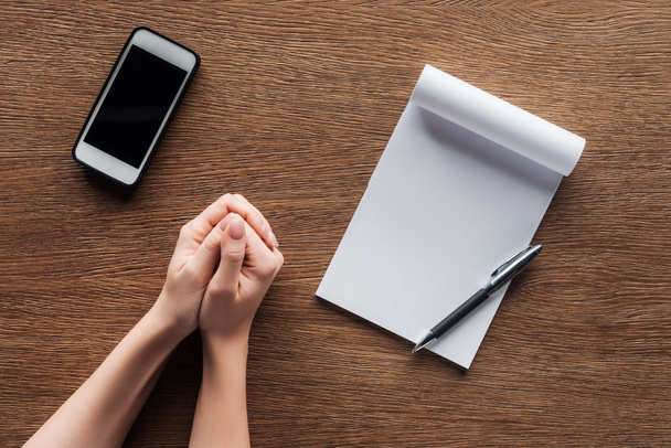 Ausgeschnittene Ansicht einer Person mit gefalteten Händen, Stift, Notizbuch mit Kopierplatz und Smartphone mit leerem Bildschirm auf Holzgrund - Foto, Bild