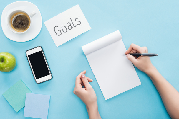 cropped view of woman writing in notebook, lettering 'goals' on card, coffee, sticky notes and smartphone with blank screen on blue background  - Photo, Image