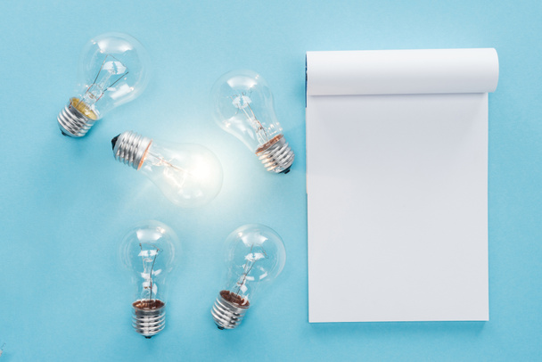 vista superior del cuaderno en blanco con bombillas sobre fondo azul, con nuevo concepto de idea
 - Foto, Imagen