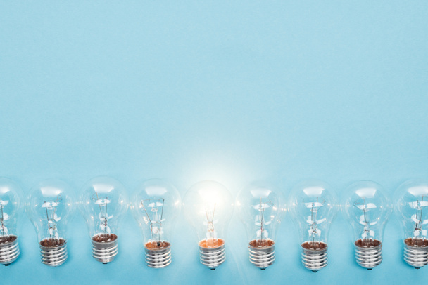 top view of light bulbs in row and one of them glowing on blue background, having new ideas concept - Photo, Image