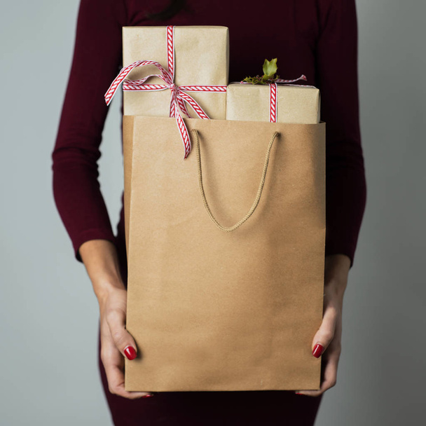 primer plano de una joven mujer caucásica sosteniendo una bolsa de compras de papel llena de regalos sobre un fondo blanquecino
 - Foto, imagen