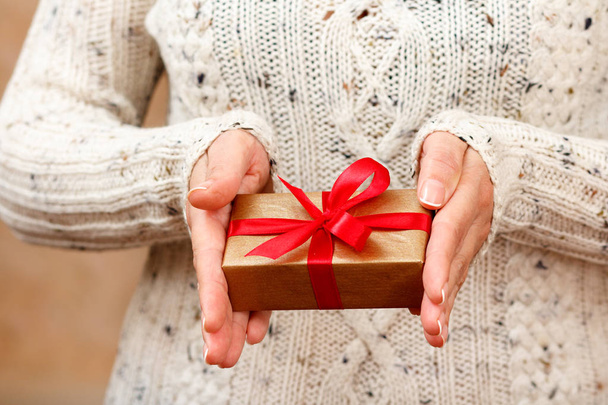 Een vrouw met een geschenkdoos met een rood lint in haar handen. Ondiepe scherptediepte, selectieve focus op de doos. Concept van het geven van een geschenk op vakantie of verjaardag. - Foto, afbeelding