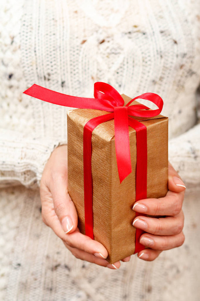 Frau mit einer Geschenkschachtel, die mit einer roten Schleife in ihren Händen gebunden ist. Geringe Schärfentiefe, selektiver Fokus auf die Box. Konzept der Bescherung im Urlaub oder zum Geburtstag. - Foto, Bild