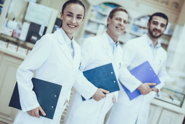 Three Pharmacists Posing. Pharmacists is a Caucasian Men and Young Woman. Persons Holding a Folders. People Wearing a Special Medical Uniform. People is Smiling. People Located in Pharmacy. - Foto, afbeelding