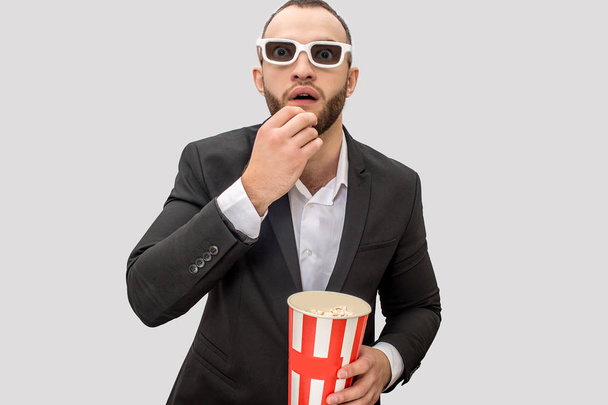 Excited young man in suit watch straight forward. He has cinema glasses on. Guy eats popcorn from small bucket. Isolated on white background. - Фото, зображення