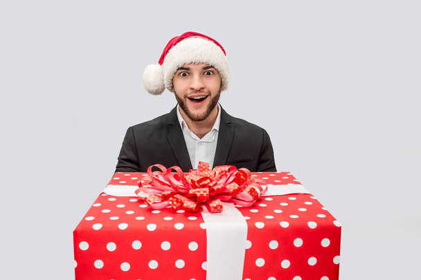 Amazed and excited young man hold big red present box. He looks on camera and smile. Guy wear Christmas hat and suit. Isolated on white background. - Foto, afbeelding