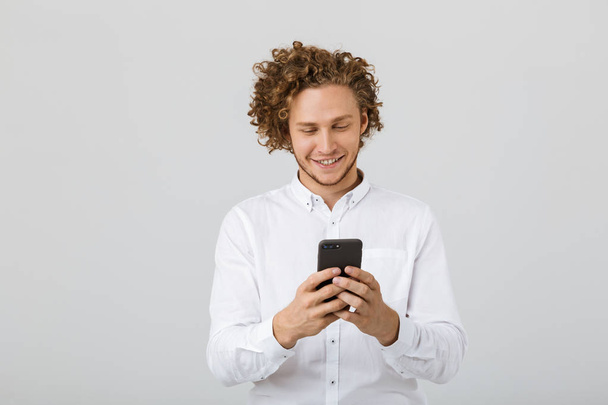 Portrait of a happy young man with curly hair isolated over white background, using mobile phone - Foto, imagen