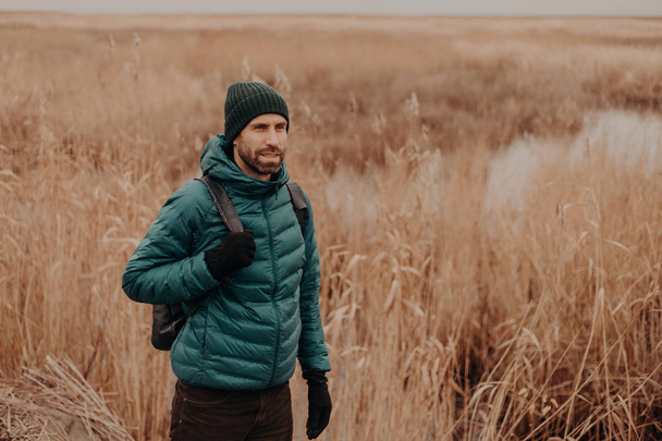 Outdoor shot of pensive male traveler dressed in warm clothes, being in good mood, has rucksack on back, walks in rural setting near field during autumn, enjoys calm atmosphere. Leisure concept - Photo, Image