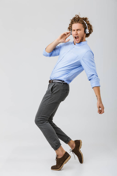 Full length portrait of a smiling young man with curly hair isolated over white background, listening to music with headphones, dancing - Foto, Imagem