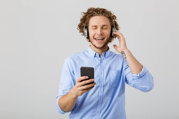 Portrait of a joyful young man with curly hair isolated over white background, listening to music with headphones, holding mobile phone - Foto, Imagem