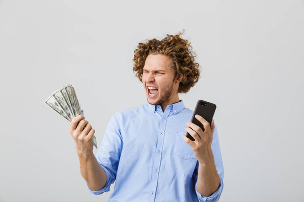 Portrait of a cheerful young man with curly hair isolated over white background, showing money banknotes, holding mobile phone - Photo, Image