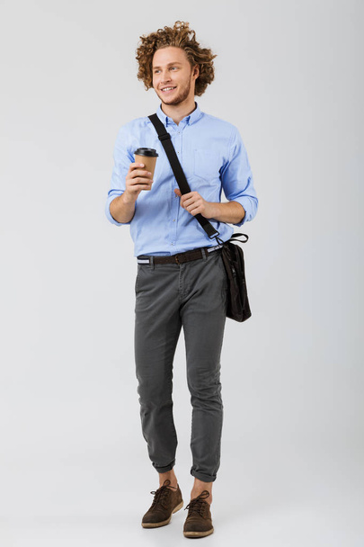 Full length of a smiling young businessman standing isolated over white background, holding takeaway coffee cup, carrying bag - Фото, изображение