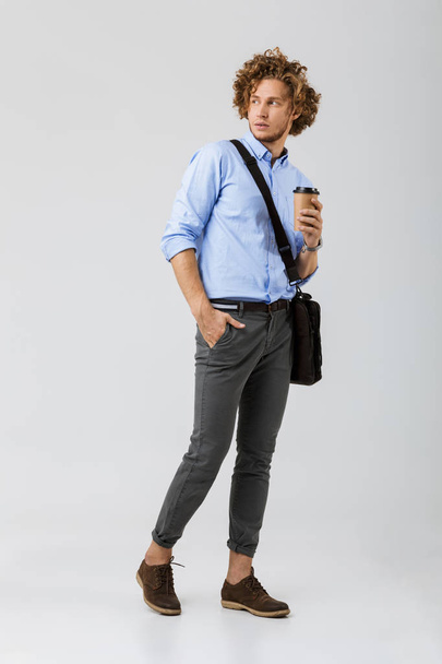 Full length of a handsome young businessman standing isolated over white background, holding takeaway coffee cup, carrying bag, looking away - 写真・画像