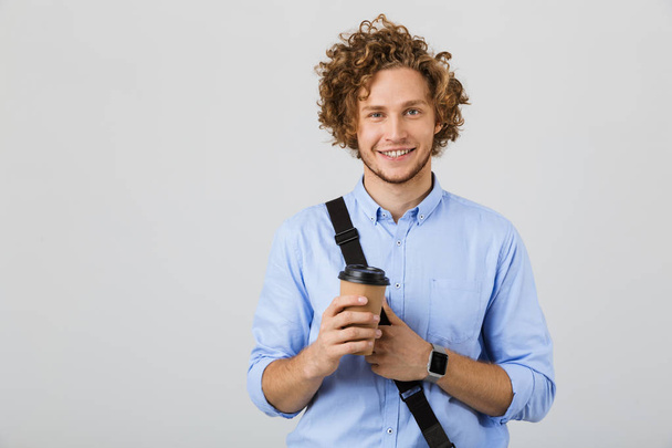 Portrait of a cheerful young man wearing shirt standing isolated over gray background, holding takeaway coffee, carrying bag - Foto, immagini