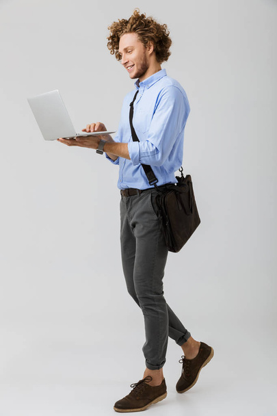 Full length of a smiling young businessman standing isolated over white background, carrying bag, working on laptop - Φωτογραφία, εικόνα